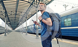 Young tourist backpacker with smartphone stand on railway station platform and waiting for train. Travel concept. Copy space