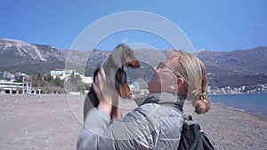 Young tourist blonde woman walks with her dachshund dog on the shore of the sea, throws the puppy up, kisses him