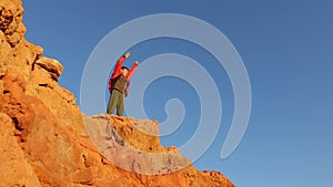 A young tourist boy climbs to the top of a mountain and raises his hand up. A tourist boy stands