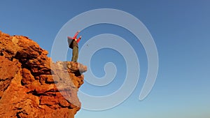 A young tourist boy climbs to the top of a mountain and raises his hand up. A tourist boy stands