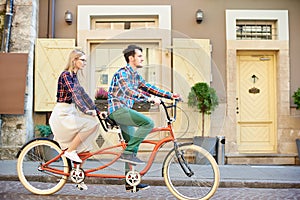 Young tourist couple, handsome man and pretty blond woman riding tandem bicycle along city street.