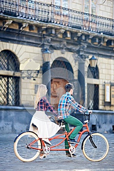 Young tourist couple, handsome man and pretty blond woman riding tandem bicycle along city street.