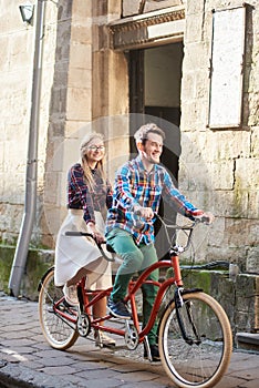 Young tourist couple, handsome man and pretty blond woman riding tandem bicycle along city street.