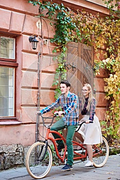 Young tourist couple, handsome man and pretty blond woman riding tandem bicycle along city street.