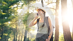 Young tourist girl backpacker in hat chatting with smartphone during walking in summer forest