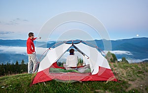 Young tourist male shows away to moon his companion female in mountains at dawn