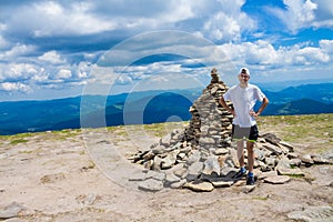 Young tourist man in a cap with hands up on the top of the mountains admires the nature