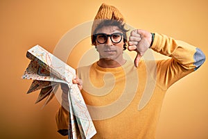 Young tourist man holding map wearing glasses and cap over isolated yellow background with angry face, negative sign showing