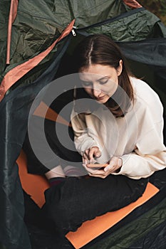 A young tourist sits in a tent in nature and looks at the phone. Surfing the Internet outside the city on hike