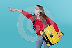 Young tourist wearing a protective mask believes COVID-19 is carrying a suitcase and points to an empty space on a blue background