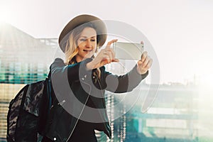 Young tourist woman in hat and with backpack stands at airport and takes photos, shoots video on smartphone`s camera.