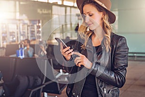 Young tourist woman in hat stands at airport, uses smartphone. Hipster girl checks email, chatting, browsing internet.