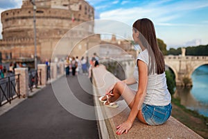 Young tourist woman sitting on the ancient embankment Tiber in Roma at sunset. Near of bridge and castle of Angels. Saint Angelo