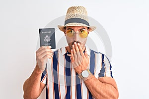 Young tourtist man wearing summer hat and holding USA passport over isolated background cover mouth with hand shocked with shame