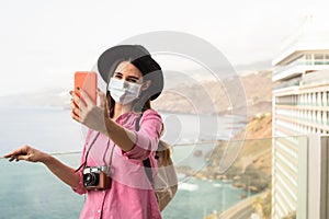 Young travel woman taking selfie with mobile smartphone wearing face surgical mask - Travel influencer having fun in vacation