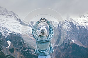 Young traveler girl in gloves standing over mountain peaks and making heart by hands