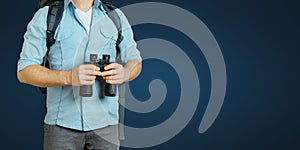 Young Traveler Man With Backpack And Binoculars Seeking Direction On Blue Background. Hiking Tourism Journey Concept