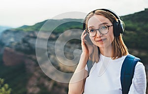 Young traveler woman with backpack listening to favorite music on wireless headphones while walking on top of mountain and enjoy