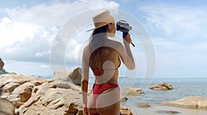 Young traveler woman films nature on camera, dressed in red swimsuit and a hat, outdoors, summertime
