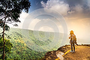 Young traveling woman with backpack hat and camera in right hand stand on the top of the mountain cliff watching beautiful view