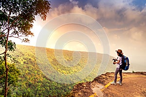 Young traveling woman with backpack hat and camera stand on the top of the mountain cliff watching beautiful view of woods and sky
