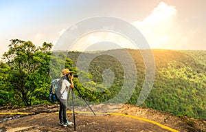 Young traveling woman with backpack hat and camera on tripod stand on the top of the mountain cliff watching beautiful view