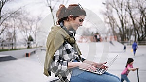 A young trendy brunette is sitting on a bench with a laptop on her knees, typing. Headphones on the neck, glasses. In