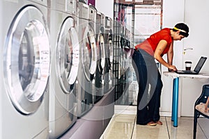 Young trendy fashion brunette use laptop computer and wait in a laundry mat service store in the city - washing clothes activity