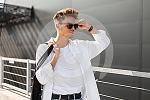 Young trendy hipster man with a stylish hairstyle straightens black sunglasses. Handsome modern guy in fashionable white clothes