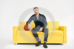 Young trendy man sitting with basketball ball on yellow sofa over white background