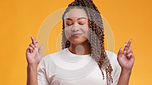 Young trustful african american lady crossing fingers and making cherished wish, orange studio background