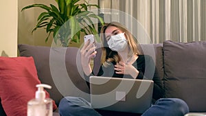 Young Turkish woman with a facial mask, having a video call on her smartphone while sitting on the sofa.