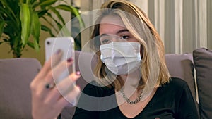 Young Turkish woman with a facial mask, having a video call on her smartphone while sitting on the sofa.