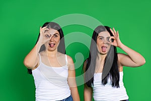 Young twin girls gesturing okay make faces with tongues out on a green background with space