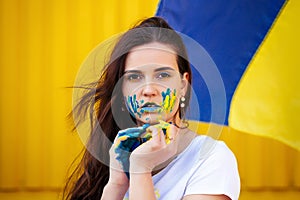 Young ukrainian girl posing with blue and yellow marks on hand and face, ukrainian flag on backgroumd