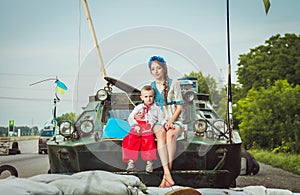 Young ukrainian woman with child boy sit on armored personnel carrier on roadblock against background of sandbags