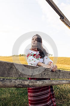 Young Ukrainian woman stands in traditional national clothes near wooden fence on meadow against sky background