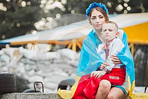 Young ukrainian woman wrapped in flag sits with child boy on roadblock against background of sandbags