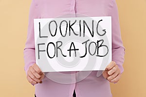 Young unemployed woman holding sign with phrase Looking For A Job on beige background, closeup