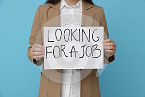 Young unemployed woman holding sign with phrase Looking For A Job on light blue background, closeup