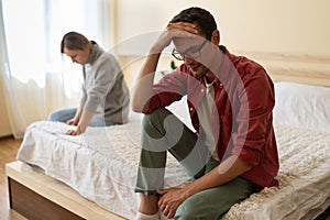 Young unhappy family couple sitting separately on bed at home, having problems in relationship