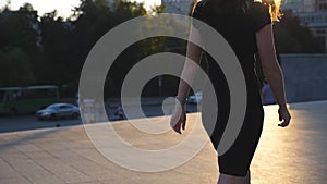 Young unrecognisable girl in black dress walking along urban street at sunset time. Attractive business woman going in