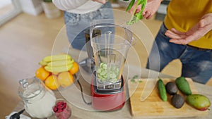 Young unrecognizable couple putting vegetables and greenery in blender cooking healthful smoothie in the morning at home