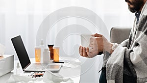 Young unrecognizable guy having fever wrapped in blanket and drinking hot tea, watching film on laptop, panorama