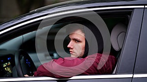 Young unshaven man in red jacket with black hood sitting at wheel of a car