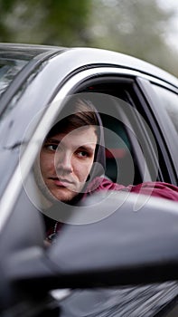 Young unshaven man in red jacket with black hood sitting at wheel of a car