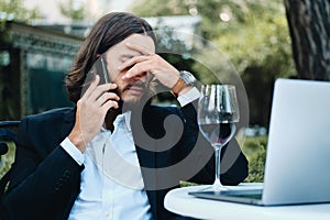 Young upset businessman with glass of wine and laptop frustratingly talking on cellphone in restaurant outdoor