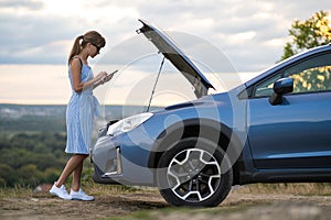 Young upset woman driver talking on mobile phone near a broken car with open hood inspecting engine having trouble with her