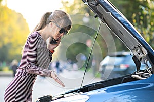 Young upset woman driver talking on mobile phone near a broken car with open hood waiting for help having trouble with her vehicle