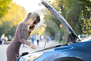 Young upset woman driver talking on mobile phone near a broken car with open hood waiting for help having trouble with her vehicle